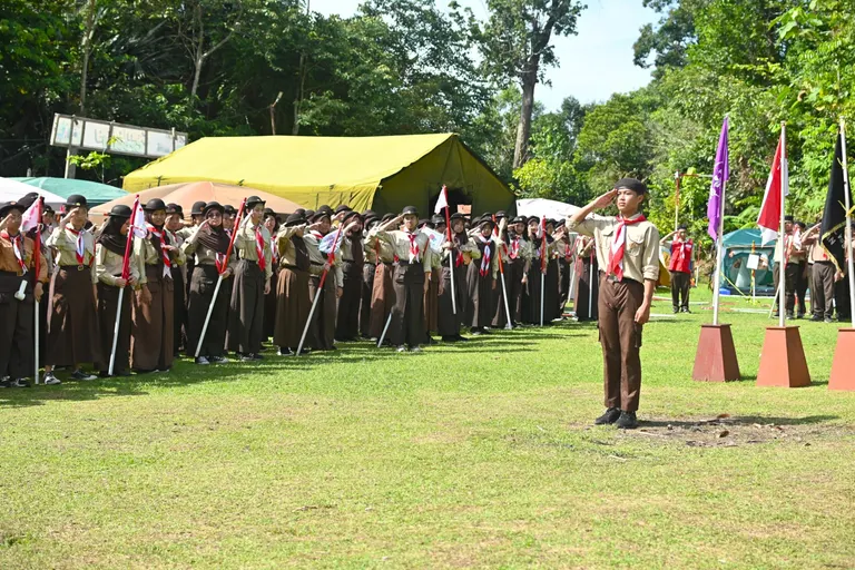 Siswa berseragam pramuka berbaris di lapangan berumput dekat tenda