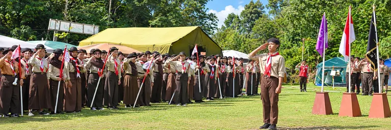 Pasukan pramuka berseragam memberi hormat di lapangan perkemahan, dengan tenda dan bendera di latar belakang